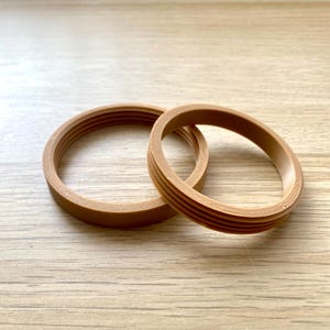 May include: Two light brown, circular objects with a ridged design. The objects are made of a solid material and are resting on a light-coloured wooden surface. The objects appear to be rings or bands.