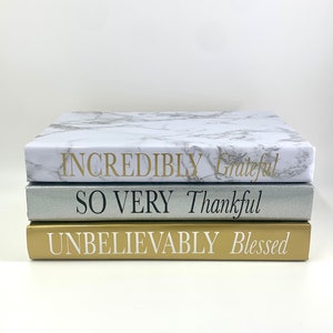 May include: A stack of three decorative books with gold, silver, and marble covers. The top book has the text "Incredibly Grateful" in gold lettering. The middle book has the text "So Very Thankful" in black lettering. The bottom book has the text "Unbelievably Blessed" in white lettering.