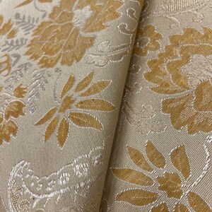 以下が含まれることがあります： ベージュの生地に、金色と銀色の花柄が施されています。生地は、織り込まれたような質感があります。