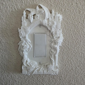 May include: White decorative light switch cover with a fantasy dragon theme. The cover features a central switch surrounded by a detailed relief of dragons, shields, and castle-like structures. The design is intricate and three-dimensional, adding a unique touch to any room.