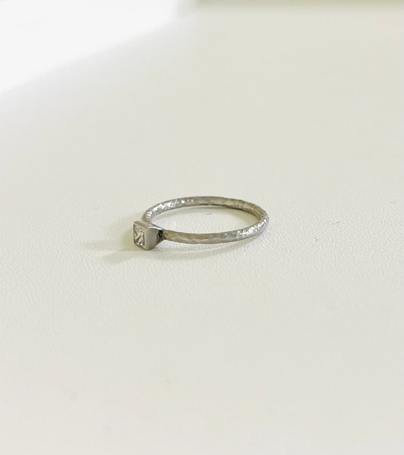 May include: A silver ring with a square-shaped clear stone set in the band. The band is textured and has a hammered finish.