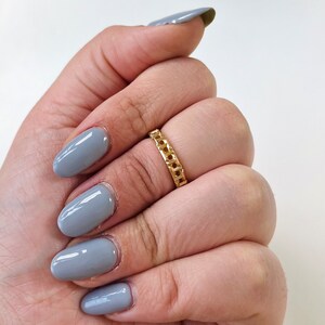 May include: A gold chain ring with a simple design. The ring is on a hand with gray nail polish.