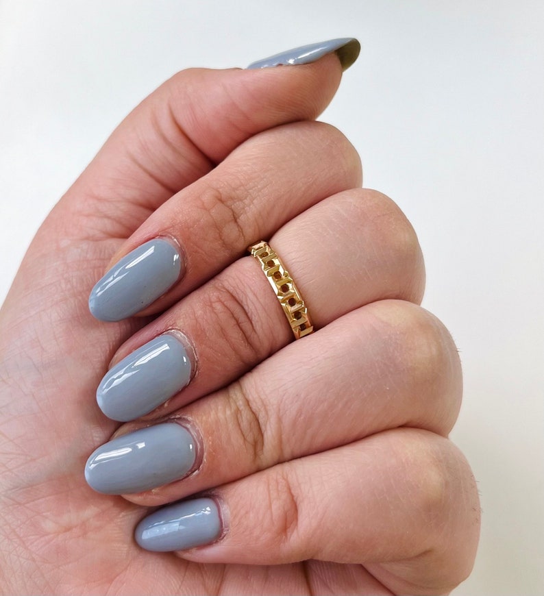 May include: A gold chain ring with a simple design. The ring is on a hand with gray nail polish.