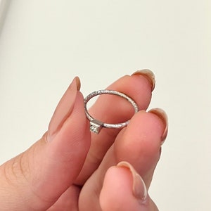 May include: A silver ring with a small, clear gemstone set in the center. The band of the ring has a textured finish.