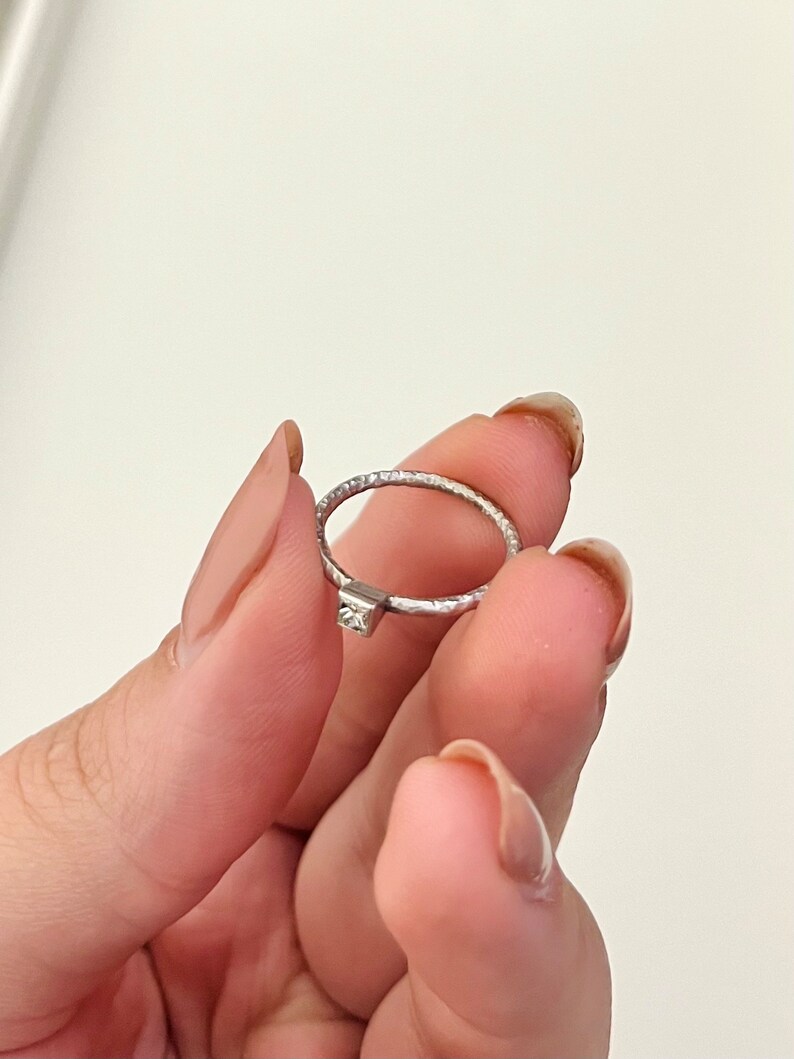 May include: A silver ring with a small, clear gemstone set in the center. The band of the ring has a textured finish.