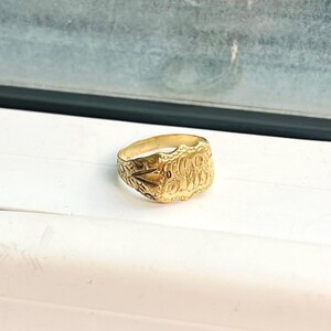 Op de afbeelding: Een gouden zegelring met een ovaalvormig vlak voorzien van een gegraveerd monogram van verstrengelde letters. De ringband en de randen van het vlak zijn versierd met gedetailleerde, textuurrijke florale krulpatronen in een glanzende afwerking.