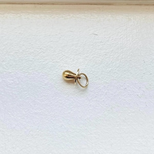 May include: A small, gold-colored pacifier charm. The charm has a rounded nipple shape and a circular ring. The charm is likely made of metal and is designed to be a pendant or charm for a necklace or bracelet.