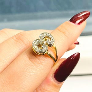 May include: A gold ring with a double-loop design, encrusted with small, clear gemstones. The ring is on a finger with a maroon nail polish.