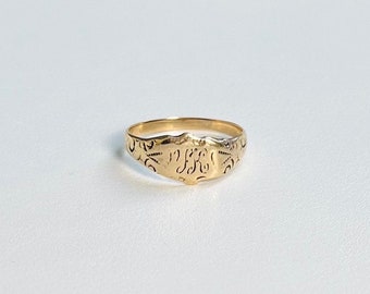Könnte beinhalten: Ein goldener Siegelring mit einem gravierten Monogramm. Der Ring hat eine einfache Band und eine erhabene ovale Oberseite mit einem detaillierten Design.