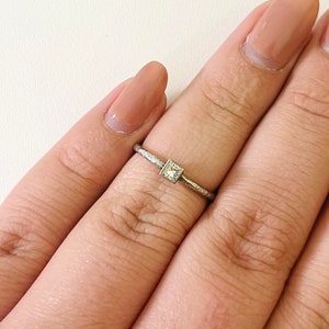 May include: A silver ring with a small, square-shaped diamond in the center. The ring is on a person's finger.