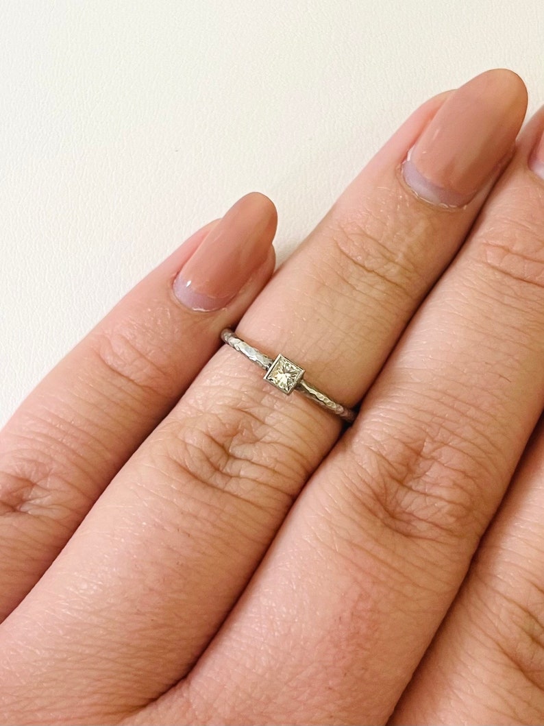 May include: A silver ring with a small, square-shaped diamond in the center. The ring is on a person's finger.