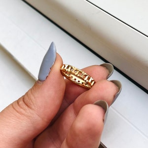 May include: A gold ring with a square-shaped design. The ring is on a finger.