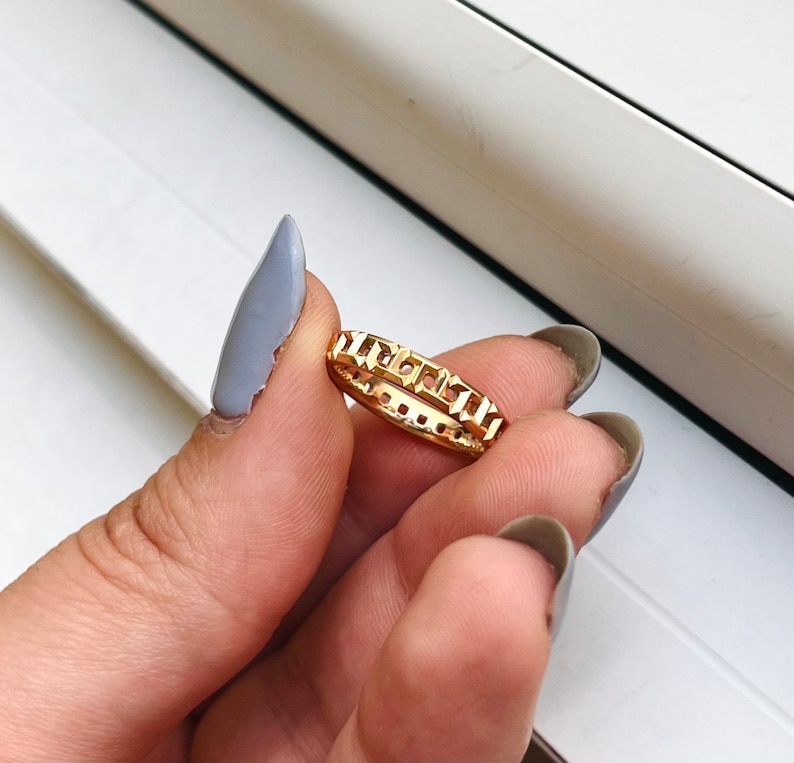 May include: A gold ring with a square-shaped design. The ring is on a finger.