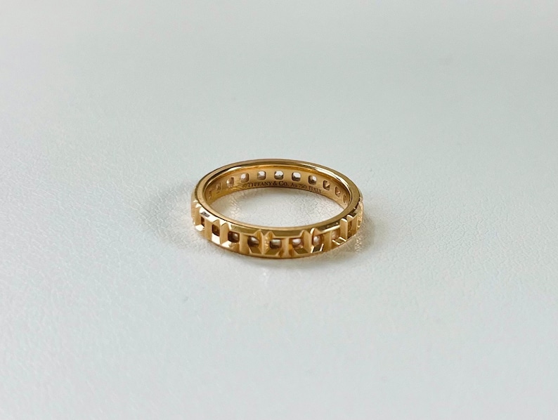 May include: A gold band ring with a repeating pattern of squares and diamonds. The ring is inscribed with "Tiffany & Co. 1837"