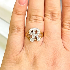 May include: A gold-coloured ring featuring the capital letter "R" set with small, clear gemstones. The ring has a split band design with a decorative cutout on each side of the letter. The ring is worn on a finger.