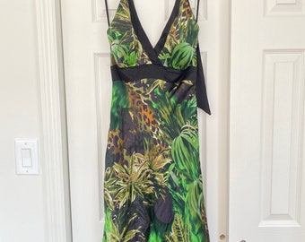 May include: A sleeveless dress with a V-neck and a black satin ribbon. The dress features a vibrant green and black tropical print with a leopard pattern. The dress is hanging on a beige hanger against a white door.
