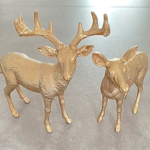 May include: Two gold-coloured deer figurines. One deer is standing with its head held high, while the other deer is standing with its head lowered. Both deer have a textured surface.