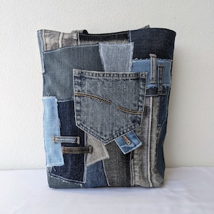 May include: A large tote bag made from recycled denim fabric. The bag is patchwork with different shades of blue denim and has a large pocket on the front.