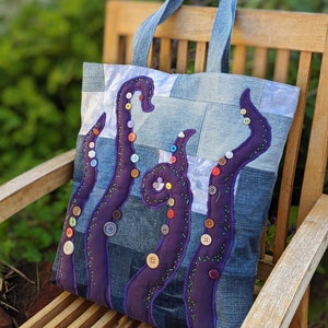 May include: A denim tote bag with a purple octopus design. The octopus is made of fabric and has colorful buttons for eyes. The bag has a patchwork design and is made from recycled denim.