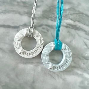 Könnte beinhalten: Zwei weiße, kreisförmige Ornamente mit maritimem Thema. Jedes Ornament zeigt geprägte Muscheln, Seesterne und Anker. Das Wort "MARINE" ist auf jedem Ornament geprägt. Eines hat eine silberne Kordel, das andere ein türkisfarbenes Band.