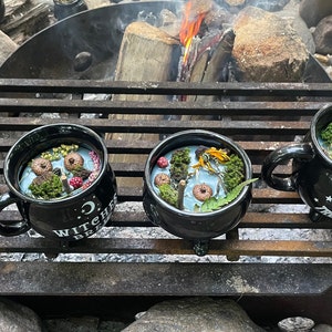 Op de afbeelding: Vier zwarte, ketelvormige bekers met handvatten, gevuld met blauwe was en decoratieve elementen, zijn gerangschikt op een zwart metalen rooster. De bekers hebben verschillende tekst aan de voorkant, waaronder "Witch's Brew" en "Toil and Trouble".