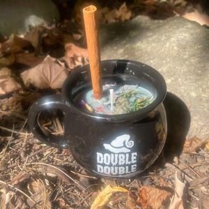 Op de afbeelding: Zwarte keramische ketelbeker met een witte heksenhoed en de tekst "Double Double". De beker is gevuld met een zwarte kaars en een kaneelstokje.