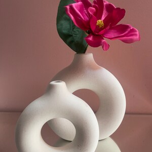 May include: Two off-white, donut-shaped vases with a pink and green flower arrangement. The flower is a vibrant magenta with a yellow center. The vases have a matte finish and are displayed on a reflective surface.
