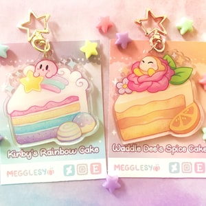 May include: Two colorful acrylic charms featuring Kirby and Waddle Dee from the video game series Kirby. The charms are shaped like slices of cake with Kirby on a rainbow cake and Waddle Dee on a spice cake. The charms are attached to gold star-shaped keychains.