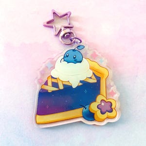 May include: A blue and purple pie slice with a yellow crust and a white whipped cream topping. A small blue blueberry character sits on top of the whipped cream. A yellow flower with a purple center and a blue blueberry are on the side of the pie slice. The charm is attached to a silver star-shaped keychain.