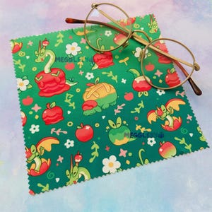 May include: A green microfiber cleaning cloth with a repeating pattern of cartoon dragons, apples, and flowers. Gold-rimmed eyeglasses rest on the cloth. The cloth has the text "MEGGLESY" in white.