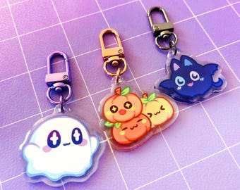 May include: Three acrylic keychains featuring cartoon characters: a white ghost with star-shaped eyes, a cluster of three orange pumpkins with expressive faces, and a dark blue bat-like creature with large eyes, each attached to a metal clasp.