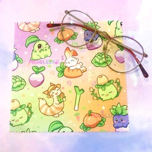 May include: Gold-rimmed eyeglasses rest on a microfiber cleaning cloth. The cloth features a colorful pattern of cartoon vegetables and creatures. The text "MEGGLESY" is printed on the cloth. The background is a gradient of yellow, pink, and purple.