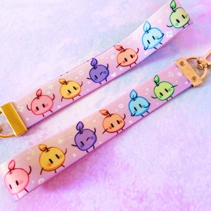 May include: Two pink wristlet keychains with a repeating pattern of cartoon fruit characters in various colors. Each keychain has a gold-colored clasp and hardware. The background is a soft, pastel gradient of pink, blue, and white.
