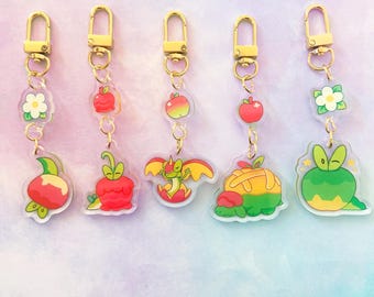 May include: Five acrylic keychains with gold-colored hardware. Each keychain features a different design, including apples, flowers, and a dragon. The keychains are attached to a gold-colored clasp.