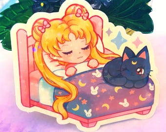 May include: A sticker featuring a cartoon illustration of a blonde-haired girl sleeping in a pink bed with a black cat. The bed has a purple and pink patterned blanket with stars, moons, and bunnies. The girl has her eyes closed and is wearing a pink and white nightgown. The cat is curled up on the bed and has a crescent moon on its forehead.