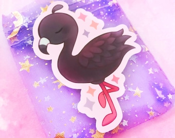 May include: A sticker featuring a cartoon flamingo in shades of black and burgundy, with a white beak and pink legs. The sticker is placed on a small, sheer purple bag decorated with gold stars and moons.