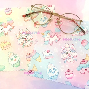 May include: Round gold-rimmed eyeglasses rest on a pastel-colored fabric. The fabric is patterned with cartoon unicorns, ice cream cones, cupcakes, and other whimsical designs. The text "MEGGLESY" is visible on the fabric.