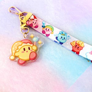 May include: A lanyard and keychain featuring a cartoon character. The lanyard is white with colorful character illustrations. The keychain is a clear acrylic charm with a yellow character wearing a hat and holding a wand. The hardware is gold-colored.