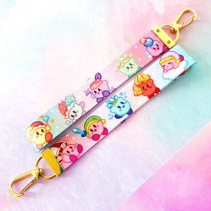 May include: Two colorful lanyards with gold-colored clasps. Each lanyard features a repeating pattern of cartoon characters in various outfits and colors, set against a pink and light blue background. The lanyards are made of a smooth, printed fabric.