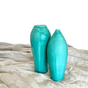 Op de afbeelding: Twee turquoise keramische vazen in verschillende maten en vormen. De vazen staan op een lichtgrijs stoffen oppervlak.