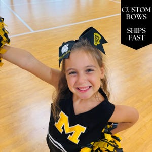 May include: A young girl smiles, wearing a black and yellow cheerleading uniform and bow. The bow has the word "CHEER" in gold glitter. The uniform has a large yellow "M" on the front. The text "CUSTOM BOWS SHIPS FAST" is in the background.