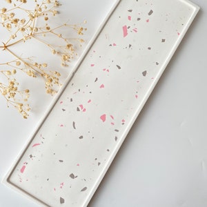 Puede incluir: Una bandeja rectangular blanca con un patrón de terrazo con motas rosas y grises. La bandeja está hecha de resina y tiene un acabado liso y pulido.
