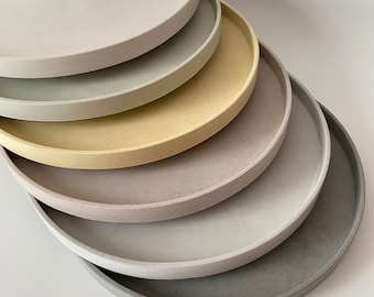May include: A set of six round concrete trays in various shades of gray, beige, and green. The trays are stacked on top of each other, with the largest tray at the bottom and the smallest tray at the top.