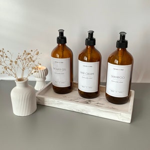 May include: Three amber glass bottles with black pump tops and white labels. The labels read "Shower Gel", "Hair Cream", and "Shampoo". The bottles are sitting on a white marble tray.
