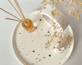 May include: A white ceramic tray with a terrazzo pattern featuring black, gold, and pink flecks. The tray has a white ceramic cutout in the shape of a crescent moon. A small glass bottle with brown liquid and wooden sticks is on the tray. A bouquet of dried white flowers is in the cutout.