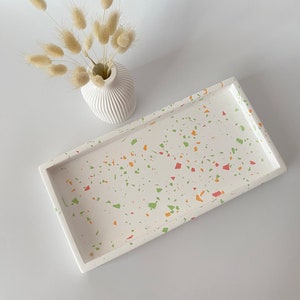 Puede incluir: Una bandeja rectangular blanca con un diseño de terrazo con motas naranjas, verdes y rojas. Un pequeño jarrón blanco con un diseño acanalado contiene flores beige secas. La bandeja está vacía y descansa sobre una superficie blanca.