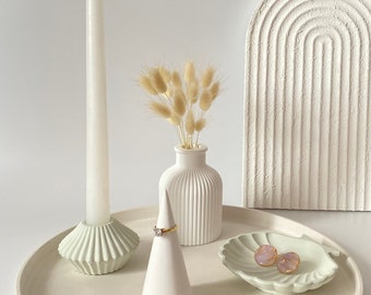 May include: A white ceramic tray with a white candle holder, a white vase with dried pampas grass, a white ring cone, and a white shell dish with two pink earrings.