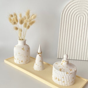 May include: A white tray with three decorative items: a vase with dried pampas grass, a ring holder, and a small container. All items have a white base with gold accents.