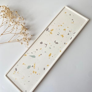 May include: A rectangular white serving tray with a terrazzo pattern in shades of yellow, gray, and green. The tray is made of resin and has a smooth, polished finish.