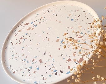May include: A white oval ceramic tray with a terrazzo pattern in shades of brown, blue, and gray. The tray is on a white surface with a sprig of dried flowers.
